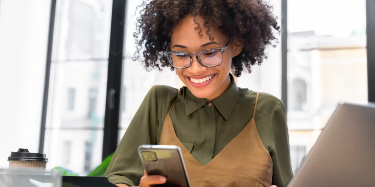 Smiling woman with glasses using a smartphone in a bright office.