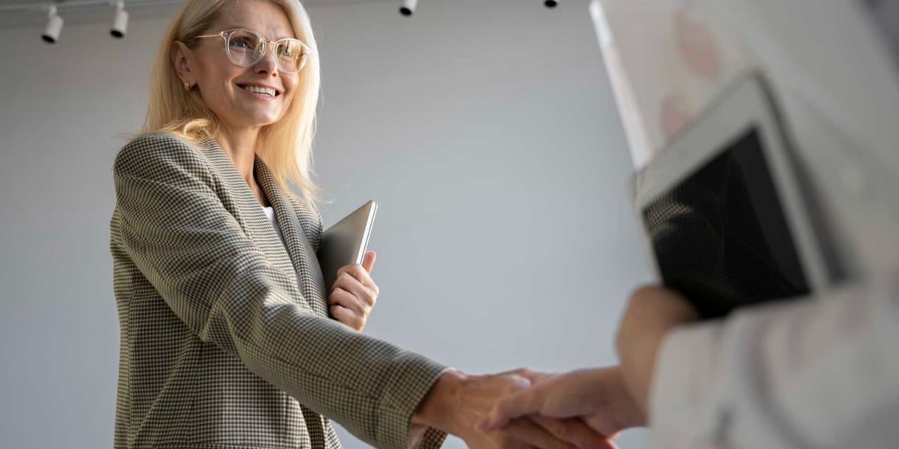 A smiling blonde-haired woman wearing glasses and a plaid blazer shakes hands with another person. She holds a silver tablet in her left arm. The other person, partially visible, is also holding a tablet. The background is light and neutral, with overhead lights. The scene conveys a professional and friendly atmosphere.