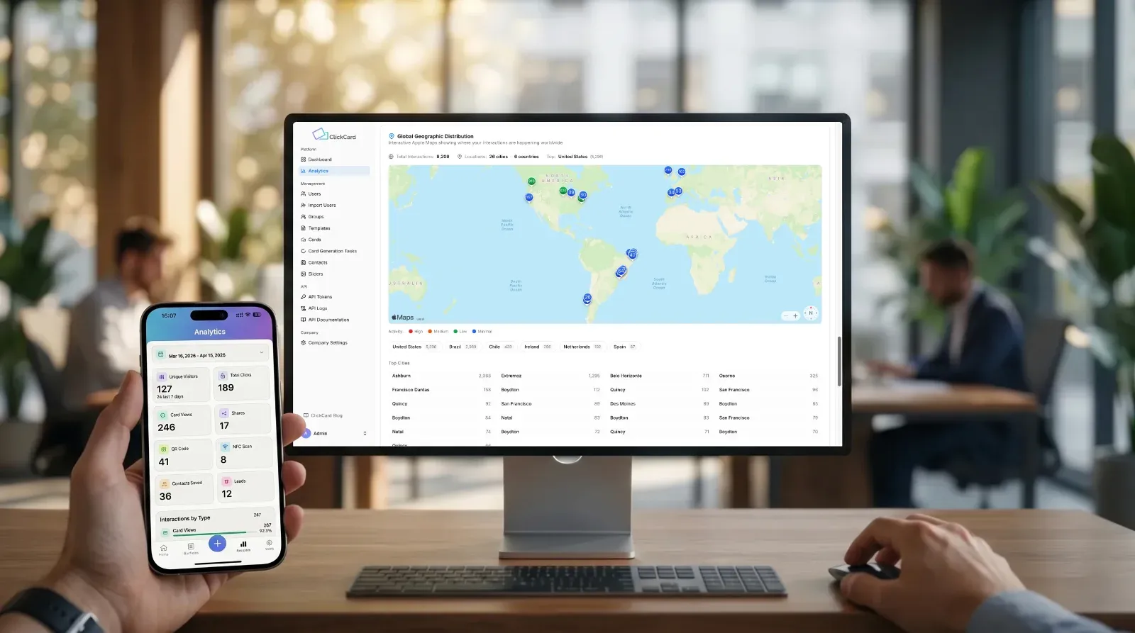 Uma visão em primeira pessoa que olha para baixo para uma mesa de madeira em um escritório de plano aberto moderno e movimentado. Uma mão no primeiro plano segura um smartphone que exibe o painel de análise do aplicativo móvel ClickCard para o período de 16 de março de 2026 a 15 de abril de 2026. A tela móvel detalha métricas principais como "Visitantes únicos" (127), "Total de cliques" (189) e "Visualizações de cartão" (246). Em frente à mão, um grande monitor de desktop exibe o painel completo de análise da web do ClickCard, com seu mapa principal de "Distribuição geográfica global". O texto na tela do monitor lista contagens de interações globais (por exemplo, Total de interações: 202) e uma lista das principais cidades. Um teclado e um mouse sem fio estão sobre a mesa. Dois colegas são visíveis trabalhando em mesas no fundo suavemente desfocado do escritório.
