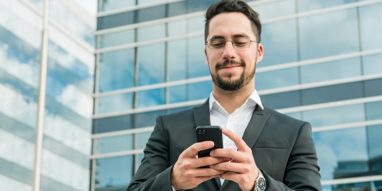 Um homem jovem, de barba e cabelos curtos, veste um terno preto com camisa branca e está usando óculos. Ele sorri levemente enquanto olha para um smartphone que segura com ambas as mãos. Ao fundo, há um prédio comercial moderno, com fachada de vidro refletindo o céu azul e algumas nuvens. A cena transmite um ambiente corporativo e profissional, com foco na tecnologia e na conectividade.