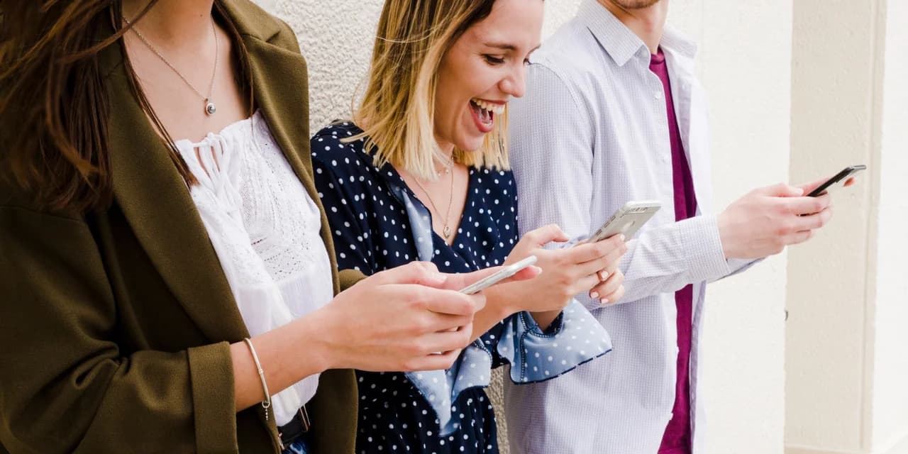 Three people are standing side by side, looking at their phones and smiling. The person in the center, with short blond hair, is wearing a navy blue blouse with white polka dots and appears to be laughing a lot. On the left is a woman with long brown hair, wearing a white lace blouse with an olive green blazer. On the right, a man in a light-colored dress shirt and a burgundy tie is holding his phone. They are standing, leaning against a light-colored wall, apparently amused by something they are seeing on their phones.